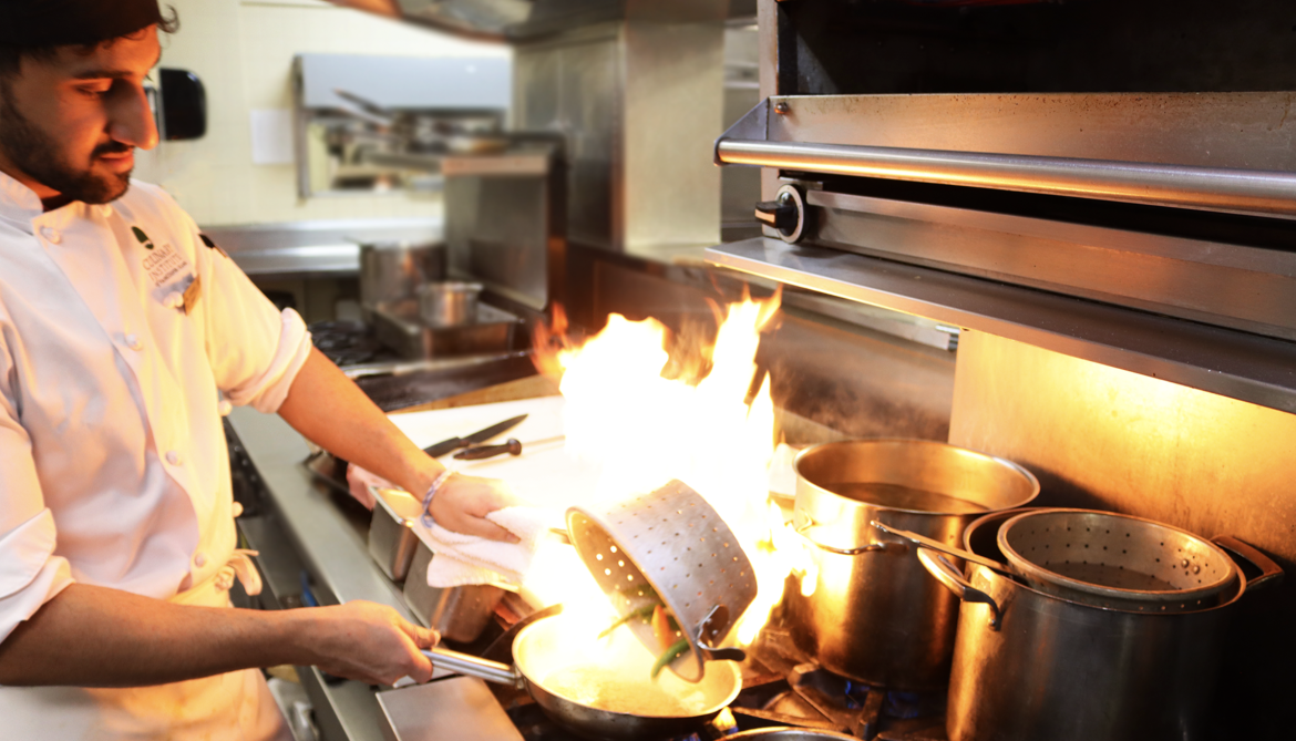 Culinary Student pouring vegetables into hot pan to sear them, creating short burst of flames in the pan