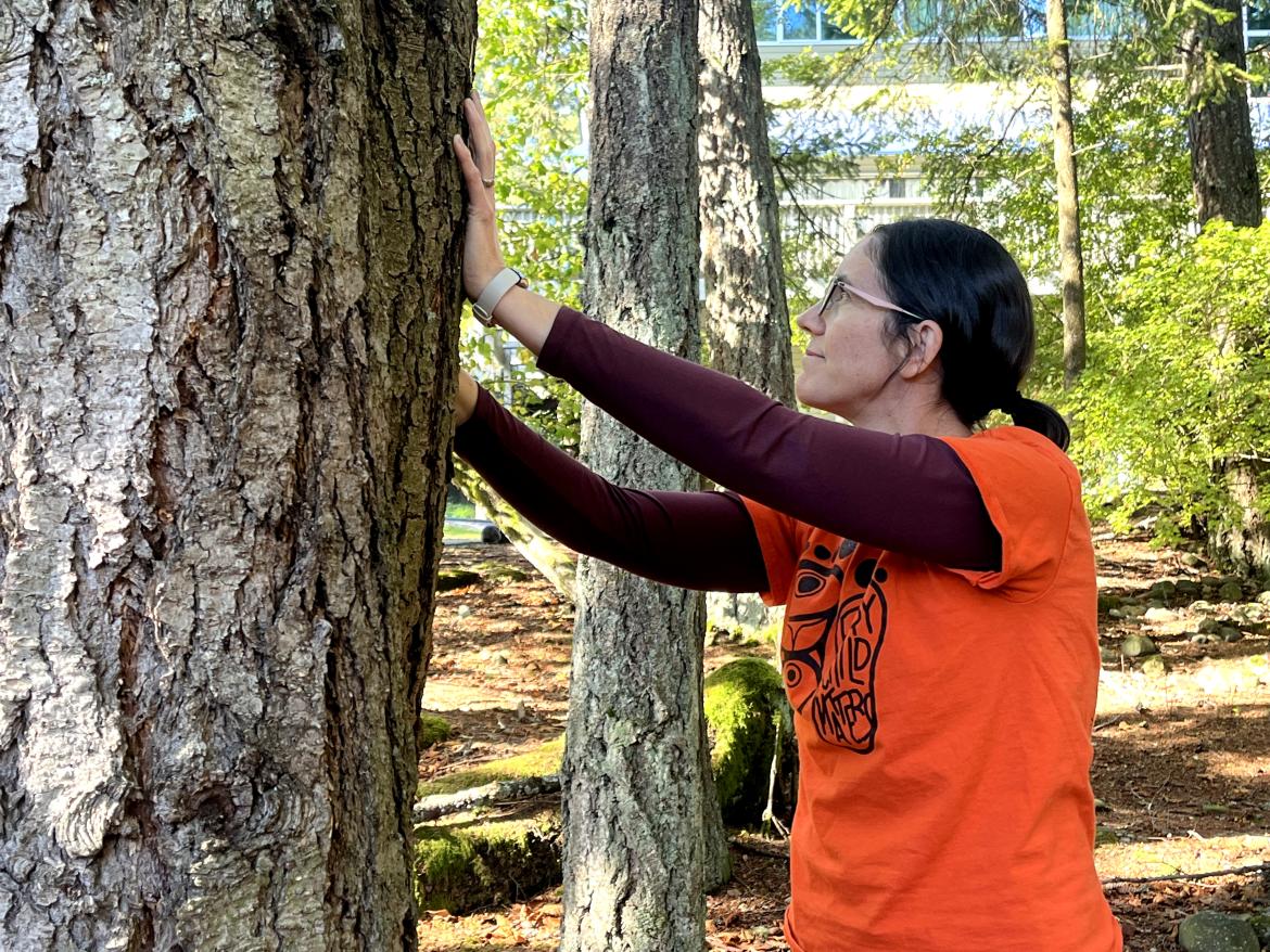 A woman wearing an orange t-shirt for Truth and Reconciliation Day putting her hands on a large tree in an act of gratitude/